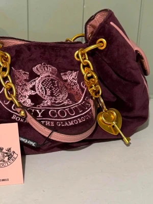 Vinröd Juicy Couture handväska - Juicy Couture handväska i vinröd sammet med rosa detaljer och guldiga kedjor. Väskan har broderad logga framtill, hjärtformat hänglås och rosa läderdetaljer. Perfekt för dig som gillar statement-accessoarer och Y2K-stil.