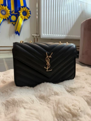 Svart YSL quiltad handväska - Snygg svart quiltad handväska från YSL med guldfärgade detaljer och kedjerem. Väskan har ett klassiskt YSL-emblem framtill och ett praktiskt innerfack med dragkedja. Tillverkad i läder med elegant V-mönster på framsidan.