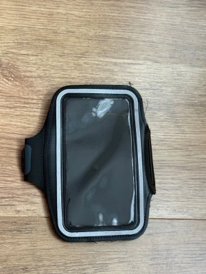 Svart sportarmband för mobil - Praktiskt svart sportarmband för mobiltelefon med genomskinligt plastfönster och justerbart kardborreband. Perfekt för träning eller löpning, tillverkat i mjukt och flexibelt material. Flera uttag för hörlurar och enkel åtkomst till skärmen.