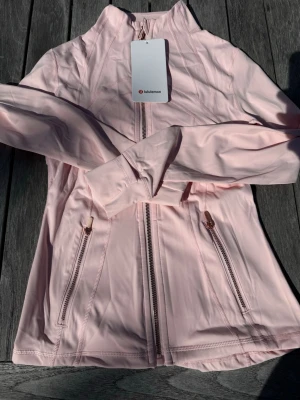 Lululemon Define Jacket – Ljusrosa (storlek 4) - Klassisk Lululemon Define Jacket i en väldigt fin ljusrosa nyans. Storlek 4 (motsvarar ca XS/S). Helt ny och aldrig använd med fin guld/metallic-logga på ryggen. Mjukt, stretchigt och fantastiskt material som sitter supersnyggt. Skickas snabbt 🚚💨 ✨ Stilren design och figursydd passform. 📩 Skicka gärna ett meddelande vid frågor eller prisförslag! 🛍️ Bundle-rabatt upp till 20%! 📦 Skickas samma eller nästa dag. 