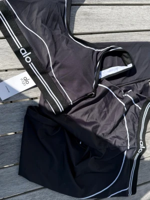 Alo Yoga Träningsset – Svart (storlek S) - Klassiskt svart Alo Yoga-set bestående av topp och leggings. Storlek S. Helt nytt och oanvänt med alla taggar kvar (NWT). Mjuk kvalitet med bra stretch och stilrena vita kontrasthänder. Skickas snabbt 🚚💨 ✨ Snygg passform som sitter perfekt. 📩 Skicka gärna ett meddelande vid frågor eller prisförslag! 🛍️ Bundle-rabatt upp till 20%! 📦 Skickas samma eller nästa dag. 