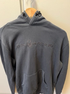 Mörkgrå hoodie från Sail Racing - Snygg mörkgrå hoodie från Sail Racing med broderad logga på bröstet. Hoodien har en klassisk känguruficka framtill, justerbar huva med snören och mjukt fleecefoder på insidan. Perfekt för dig som gillar en stilren och sportig look.