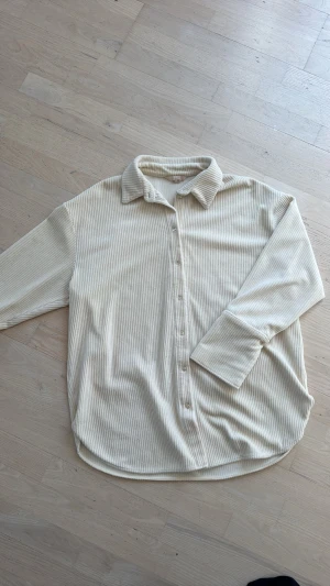 Beige ribbad manchesterskjorta - Säljer en beige långärmad skjorta i mjuk manchester med ribbad struktur. Skjortan har klassisk krage, knappar framtill och en avslappnad passform. Perfekt för dig som gillar en chill och stilren look.