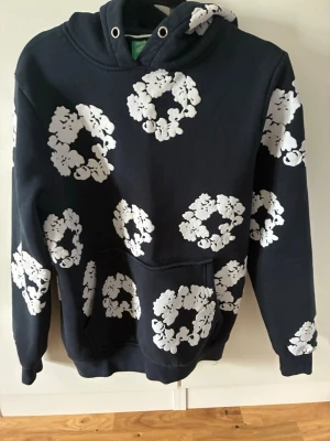 Svart hoodie med vita blommönster - Cool svart hoodie med stora vita blommönster över hela plagget. Klassisk känguruficka framtill och huva med dragsko. Perfekt för dig som vill sticka ut med en unik och trendig stil. Mjuk bomullskänsla och avslappnad passform.