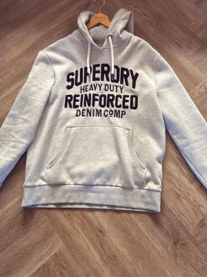 Grå hoodie från Superdry - Snygg grå hoodie från Superdry med svart texttryck framtill och känguruficka. Klassisk huva med vita dragsnören och ribbade muddar. Perfekt för en chill och avslappnad stil. Materialet känns mjukt och bekvämt.storlek xl men passar även L och M 