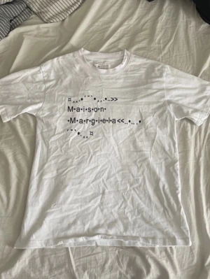 Maison Margiela vit t-shirt med tryck - Vit t-shirt från Maison Margiela med svart grafiskt tryck och text på bröstet. Klassisk rund halsringning och rak passform. Tillverkad i mjukt bomullsmaterial som känns skönt mot huden. Perfekt för en clean och modern stil.