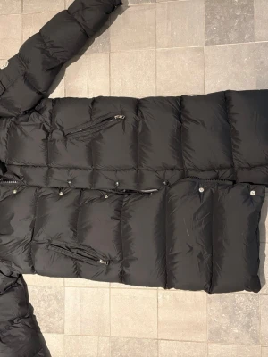 Svart lång dunjacka från Moncler - Säljer en svart lång dunjacka från Moncler med klassisk quiltad design. Jackan har flera dragkedjeförsedda fickor och tryckknappar med Moncler-logga. Perfekt för kalla vinterdagar och riktigt snygg till alla outfits.