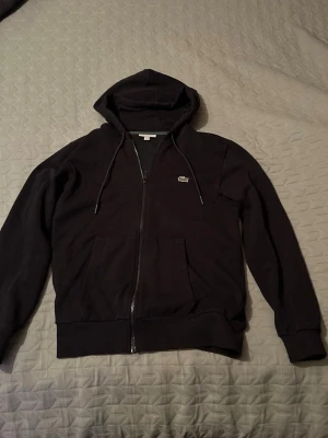 Svart zip-hoodie från Lacoste - Snygg svart hoodie från Lacoste med dragkedja framtill och klassisk krokodil-logga på bröstet. Hoodien har två fickor fram, justerbar huva med snören och ribbade muddar vid ärmar och nederkant. Perfekt för en chill och stilren look.
