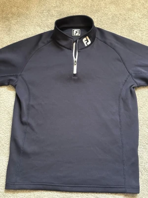 Mörkblå ziptröja från FootJoy - Mörkblå ziptröja från FootJoy med hög krage och vit dragkedja framtill. Tröjan har diskret logga på kragen och dragkedjan, samt är tillverkad i ett stretchigt funktionsmaterial. Perfekt för lager-på-lager och sportiga outfits.