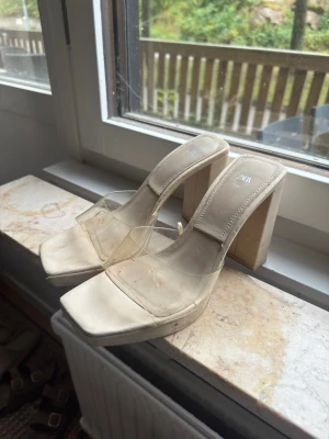 Beige klackar med transparent rem - Snygga beige pumps från Zara med fyrkantig tå och bred klack. Skorna har en transparent rem över foten och en chunky klack som ger stabilitet. Perfekta för dig som vill ha en modern och trendig look. Materialet är syntet och plast. Lite fläckar som går bort med wipes