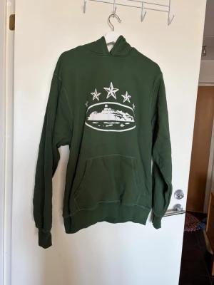 Grön hoodie Corteiz - Säljer en grön hoodie med stor ficka framtill och vit tryckt design. 