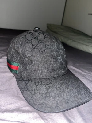 Svart Gucci keps med GG-mönster - Snygg svart keps från Gucci med klassiskt GG-monogram över hela kepsen. På sidan finns en grön och röd rand som ger en ikonisk touch. Kepsen har böjd skärm och är tillverkad i ett slitstarkt tygmaterial.