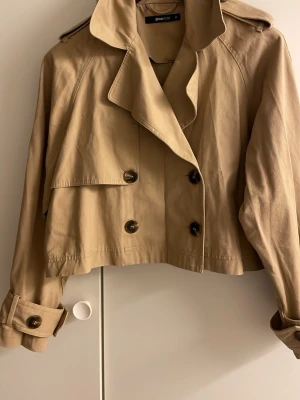 Kort beige trenchcoat från Gina Tricot - Stilren kort trenchcoat i beige från Gina Tricot. Jackan har dubbelknäppning, bred krage, axelklaffar och dekorativa knappar vid ärmsluten. Tillverkad i ett lätt tyg som ger en klassisk look. Perfekt för dig som gillar trendiga ytterplagg.