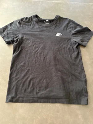 Svart Nike T-shirt i bomull - Svart klassisk T-shirt från Nike med liten vit logga på bröstet. T-shirten är i mjuk bomull och har en enkel, rak passform med rund hals och korta ärmar. Perfekt för en avslappnad och sportig stil.