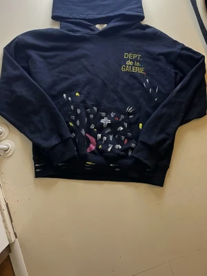 Mörkblå hoodie med tryck - Säljer en mörkblå hoodie med huva och ficka framtill. På bröstet står det 'DÉPT. de la GALERIE' i gult och grönt, och på fickan finns färgglada penseldrag i vitt, gult, rött och rosa. Hoodien har ribbade muddar och en avslappnad passform.