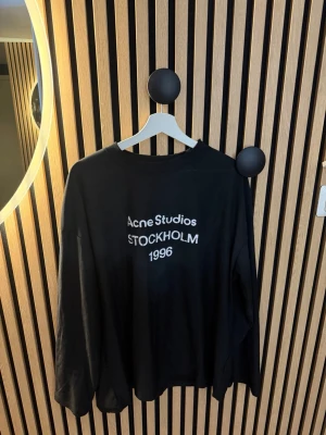 Svart långärmad topp Acne Studios - Svart långärmad topp från Acne Studios med tryck i vitt på bröstet där det står 'Acne Studios STOCKHOLM 1996'. Avslappnad passform och rund halsringning. Perfekt för en cool och enkel stil.