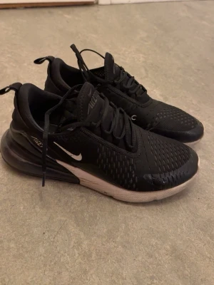 Svarta Nike Air Max 270 sneakers - Snygga svarta Nike Air Max 270 sneakers med vit sula och synlig Air-enhet i hälen. Skorna har mesh-överdel för bra ventilation, klassisk Nike-logga på sidan och dragflik bak. Perfekta för dig som gillar sportig och modern stil.