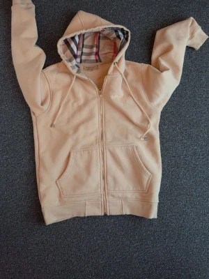 Beige hoodie med dragkedja och huva - Snygg beige hoodie med dragkedja och klassiskt rutigt foder i huvan. Hoodien har två stora fickor framtill, dragsko i huvan och ribbade muddar. Perfekt för en chill och avslappnad stil. Materialet ser ut att vara mjukt och bekvämt, troligen bomullsblandning.