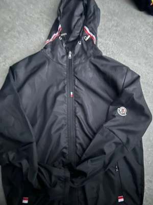 Moncler windbreaker  - Snygg svart vindjacka med huva och dragkedja framtill. Jackan har vita och röda detaljer på huvan och vid fickorna samt en broderad logotyp på ärmen. Perfekt för blåsiga dagar och enkel att matcha med olika outfits.