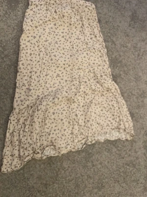 Lång blommig kjol i ljus beige - Söt långkjol i ljus beige med små blommiga mönster. Kjolen har volangdetaljer och är tillverkad i ett lätt och luftigt material, perfekt för varmare dagar.