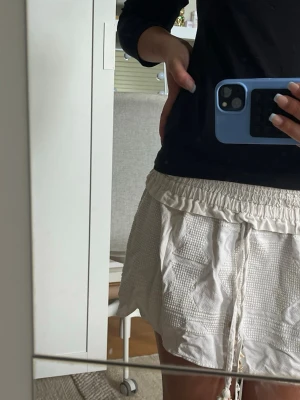 Vit kjol med snörning och struktur - Säljer en vit/beige kjol med snygg struktur och resår i midjan. Shortsen har snörning framtill och är tillverkade i ett lätt och luftigt material, perfekta för varma dagar. Passformen är loose och de har en avslappnad vibe.