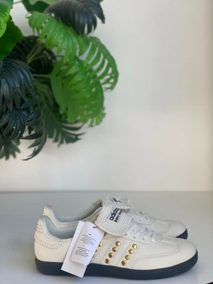 Vita Adidas sneakers med gulddetaljer - Snygga vita Adidas sneakers i läder med svarta sulor och guldnitar längs de klassiska tre ränderna. Skorna har snörning och kardborreband upptill samt diskret Adidas-logga på plösen. Perfekta för dig som vill sticka ut med coola detaljer.