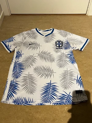 Vit Brasilien fotbollströja med bladmotiv - Snygg vit fotbollströja med blå och grå palmbladsmönster, Brasilien-emblem på bröstet och blå detaljer vid krage och ärmslut. Perfekt för dig som vill sticka ut på planen eller på läktaren. Tillverkad i lätt och ventilerande material.