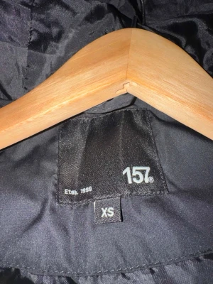 Svart lång dunjacka från 157, XS - Snygg svart lång dunjacka från 157 i storlek XS. Jackan har hög krage, raka kanaler och stängs med dragkedja och knappar. Perfekt för kalla vinterdagar och ger en stilren look.