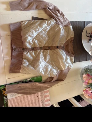 Beige quiltad kofta med knappar - Snygg beige kofta med quiltad framsida och stickade ärmar samt rygg. Koftan har ribbade muddar, rund halsringning och knäppning framtill med stora knappar. Materialmix av glansigt tyg framtill och mjuk stickning på övriga delar.