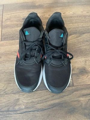 Svarta Adidas sneakers med röda ränder - Säljer ett par svarta Adidas sneakers med klassiska röda ränder på sidan och vit sula. Skorna har snörning och är tillverkade i mesh och syntetmaterial för en sportig look. Perfekta för dig som gillar stilrena och bekväma sneakers.
