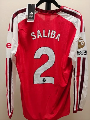 Arsena spelar version hemma matchtröja Saliba #2 - Röd och vit långärmad Arsenal matchtröja från Adidas med Saliba och nummer 2 på ryggen. Tröjan har Premier League-märke på ärmen, klubbmärke på bröstet och sponsortryck framtill. Tillverkad i lätt och ventilerande material, perfekt för fotbollsträning eller match.