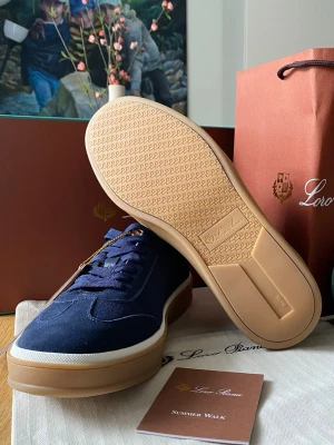 Blå sneakers från Loro Piana - Snygga sneakers i marinblå mocka från Loro Piana med vita detaljer och ljusbruna gummisulor. Klassisk låg modell med matchande blå skosnören och diskret logga på sidan. Perfekta för en stilren och avslappnad look.