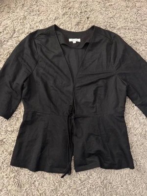 Svart omlottblus med knytband - Stilren svart blus med knytband i midjan och trekvartsärm. Blusen har en lätt v-ringad halsringning och är tillverkad i ett mjukt, vävt tyg som ger en snygg passform. Perfekt att matcha med jeans eller kjol för en trendig look. Använd fåtal gånger 
