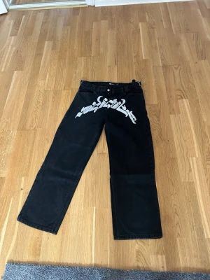 Svarta jeans med vit graffiti - Säljer ett par svarta jeans från Infinity med bred passform och vit graffiti-inspirerad text över framsidan. Jeansen har klassisk femficksdesign, bälteshällor och metallknapp. Materialet är kraftig denim som ger en cool streetwear-look.