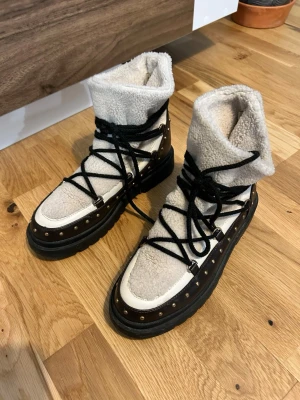 Fodrade vita boots med nitar - Säljer ett par vita moon boots med svart sula och svarta snören. Bootsens ovandel är i mjukt teddyfoder och har dekorativa nitar längs kanten. Perfekta för kalla dagar när du vill ha både stil och värme.