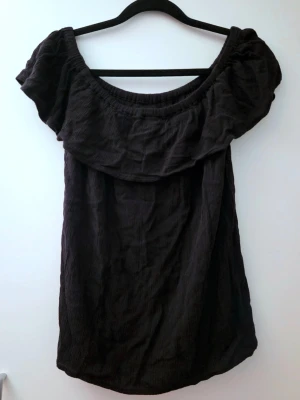 Svart offshoulder topp - Svart topp med offshoulder-modell och bred volang runt axlarna. Toppen har en loose passform och är perfekt för varma dagar. Enkel och stilren design som är lätt att matcha med accessoarer.