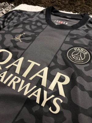 PSG x Jordan svart matchtröja - Snygg svart PSG matchtröja i samarbete med Jordan. Tröjan har ett unikt mönster i olika nyanser av svart och grått, PSG-logga på bröstet och Jordan-logga i silver. Tillverkad i lätt och ventilerande material, perfekt för fotboll eller träning.