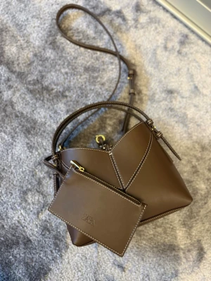 Brun handväska med axelrem från Zara - Snygg brun handväska från Zara i slätt konstläder med vita kontrastsömmar. Väskan har både handtag och justerbar axelrem samt en matchande liten clutch med dragkedja. Guldiga metalldetaljer ger en extra touch. Perfekt för dig som gillar stilrena accessoarer.