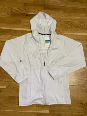 Vit vindjacka med huva från Nike - Snygg vit vindjacka från Nike med dragkedja framtill, huva och två sidofickor. Jackan har elastiska muddar vid ärmsluten och ett diskret Nike-logga på bröstet. Tillverkad i lätt och tunt material som andas, perfekt för blåsiga dagar.