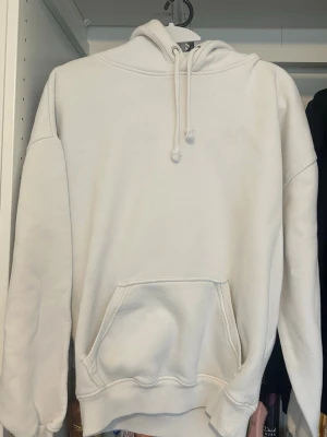 Vit hoodie med magficka och huva - En stilren vit hoodie med stor magficka och justerbar huva med dragsko. Hoodien har ribbade muddar vid ärmslut och nederkant. Perfekt för en avslappnad och bekväm look. Materialet ser ut att vara mjukt och fleece-liknande på insidan.