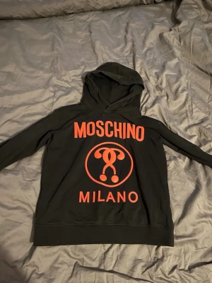 Svart Moschino hoodie med röd logga - Snygg svart hoodie från Moschino med stor röd logga och Milano-tryck på framsidan. Klassisk huva och långa ärmar. Perfekt för dig som vill sticka ut med en trendig och bekväm streetwear-look.