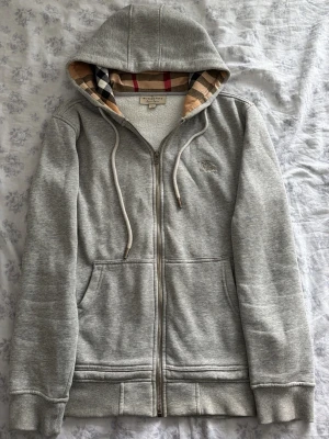 Burberry hoodie - Stilren grå Burberry hoodie med klassiskt rutigt foder i huvan och dragkedja framtill. Hoodien har fickor på sidorna, broderad logga på bröstet och justerbar huva med snören. Perfekt för en chill och stilren look. Hoodien är i strl: M. Hör av dig vid frågar! Fraktar inom 24h efter köp📦