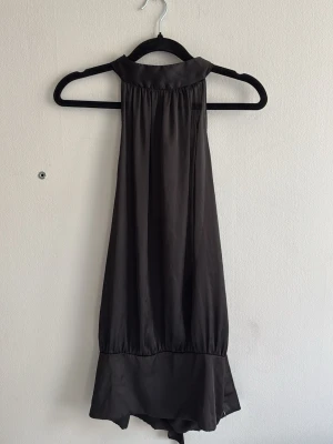Svart halterneck topp med knyt - Säljer en svart halterneck topp i ett glansigt material med bred kant nedtill och knyt i nacken. Toppen är ärmlös och har en lös passform som ger en snygg siluett. Perfekt för dig som gillar stilrena och trendiga plagg.
