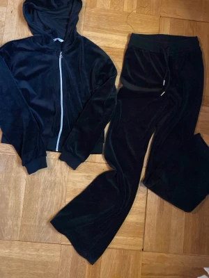Svart velourset från Ellos - Mysigt svart loungewear-set från Ellos i mjuk velour. Setet består av en hoodie med dragkedja och huva samt matchande byxor med resår i midjan och dragsko. Perfekt för chill dagar hemma eller när du vill vara extra bekväm.