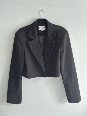 Svart cropped kavaj från NA-KD - Stilren svart cropped kavaj från NA-KD med klassisk krage och en knapp framtill. Kavajen har lång ärm och är tillverkad i ett slätt tyg som ger en snygg och modern look. Perfekt för att lyfta vilken outfit som helst.