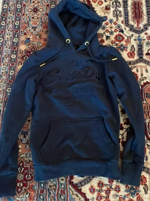 Mörkblå hoodie från Superdry - Snygg mörkblå hoodie från Superdry med broderad logga på bröstet och känguruficka framtill. Hoodien har dragsko med gulddetaljer och en klassisk huva. Perfekt för en avslappnad och trendig stil.