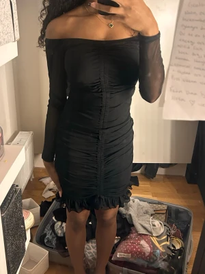 Svart offshoulder kort klänning - Snygg svart kort klänning med offshoulder-design och långa meshärmar. Klänningen har rynkade detaljer framtill och volangkant nedtill för en extra touch. Figurnära passform som framhäver siluetten.