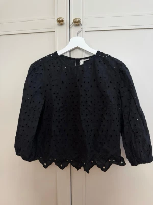 Svart broderad blus med hålmönster - Snygg svart blus med broderade hålmönster och vågig kant nertill. Blusen har rund halsringning och trekvartsärm med lätt puff. 