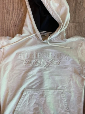 Vit Burberry hoodie med brodyr - Säljer en vit hoodie från Burberry med broderad text 'Burberry London England' på bröstet. Hoodien har en klassisk huva med dragsko, magficka och svarta detaljer på insidan av huvan. Perfekt för dig som gillar exklusiv streetwear.