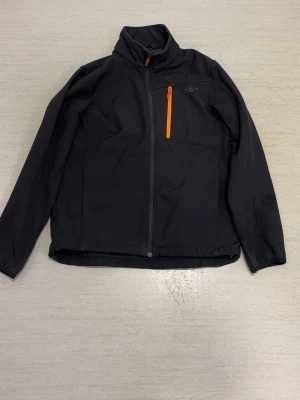 Svart vindjacka med orange dragkedja - Snygg svart vindjacka med hög krage och hel dragkedja framtill. Jackan har en orange bröstficka med dragkedja som sticker ut och ger en sportig look. Insidan är fodrad med mjuk fleece för extra komfort. Perfekt för blåsiga dagar.
